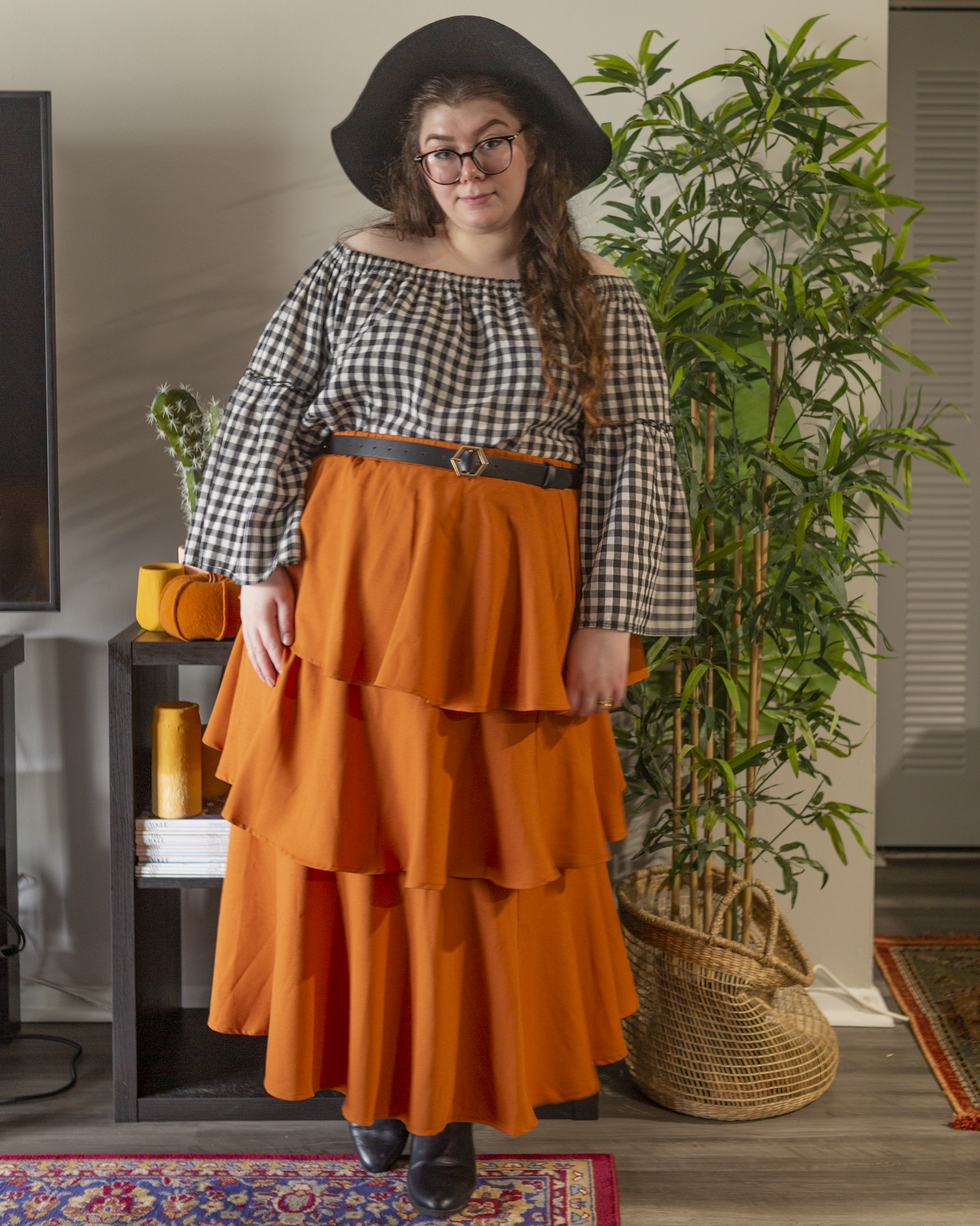 An outfit consisting of an a black wide brim floppy hat, a white and black gingham off the shoulder blouse with bell sleeves, tucked into an cinnamon orange three tier maxi skirt and black Chelsea boots.