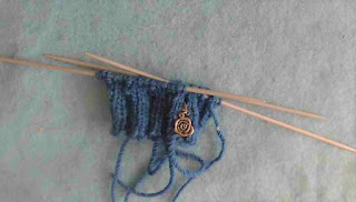 The cuff of a sock done in 2 by 2 ribbing on Double-Pointed Needles.  They're knit in blue worsted-weight yarn, and there's a silver rose stitch marker clipped into the knitting.