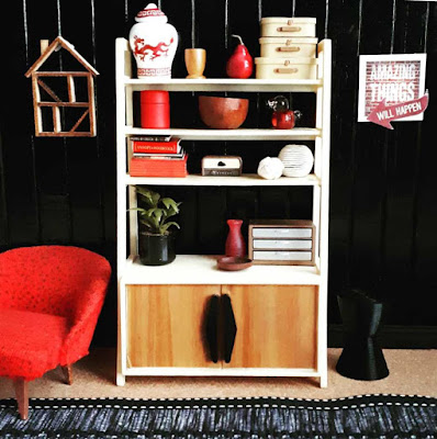 One-twelfth scale modern miniature scene with a mid-century modern cupboard unit and chair. On the unit is a variety of decorative objects including storage drawers, books, vases and a pot plant. On the wall is a picture with the words 'Amazing things will happen' and a dolls' house shadow box.
