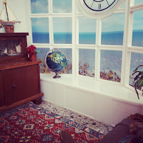 1/12 scale miniature living room scene with an antique cupboard, afghan rug and sea chest. The windows have a view over the sea, and on the windowsill stands a globe.