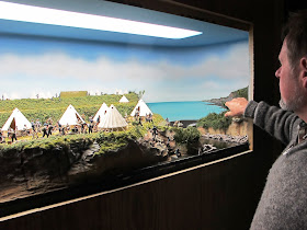 A man poses in front of a diorama of an encampment of 19th-century soldiers by the sea.