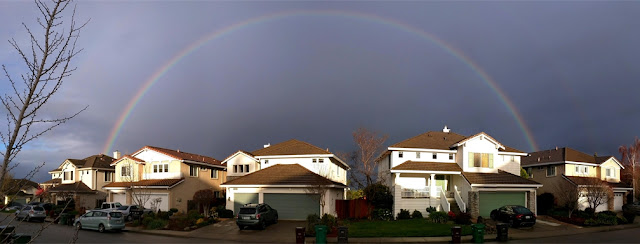 Full rainbow above neighborhood houses against an overcast sky. Garbage bins are in front of the houses.