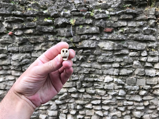 A picture of a hand holding a small, ceramic skull, Skulferatu #51, with an old, stone wall in the background.  Photo by Kevin Nosferatu for the Skulferatu Project.