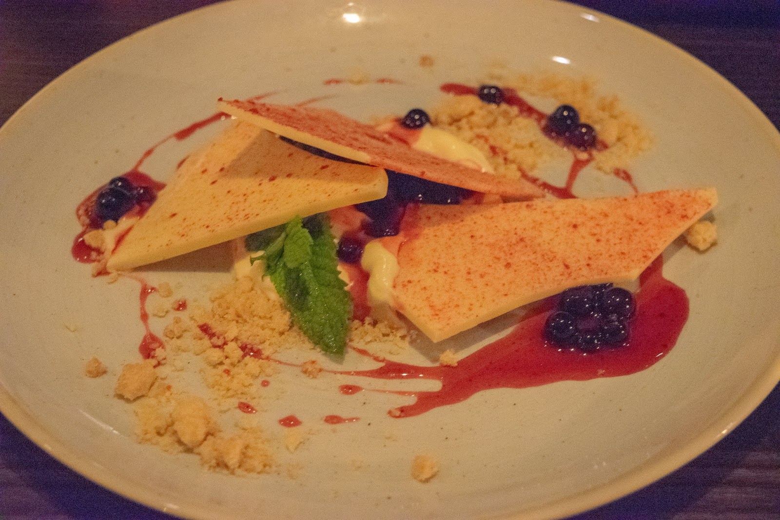 A deconstructed cheese cake with slabs of white chocolate, crumble, blackberries and red fruit sauce covering the chocolate.