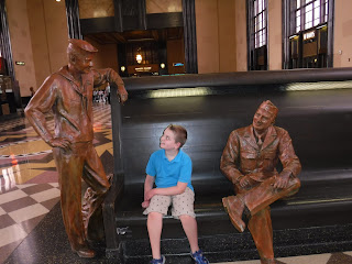 A boy sits between two life-sized statues of WWII soldiers and pretends to chat