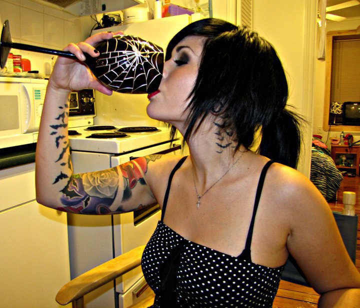 Chica gotica bebiendo de una copa con telaraña y lleva tatuajes de murcielagos por el cuello y por sus brazos