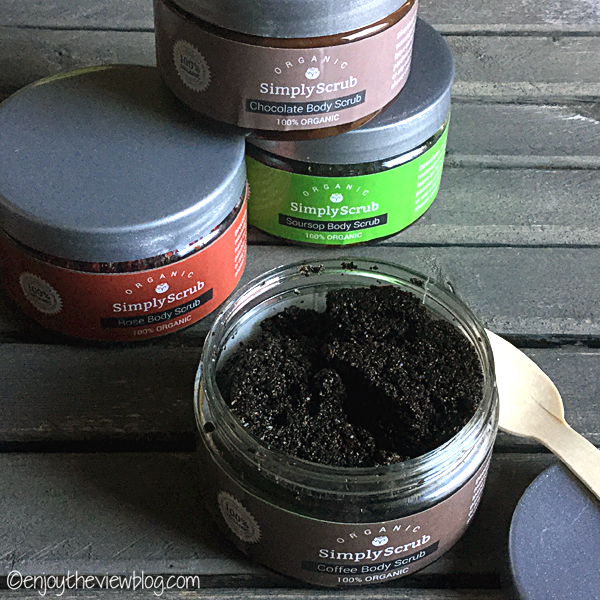 four jars of Simply Scrub Organic Body Scrubs (one open) sitting on a wooden table