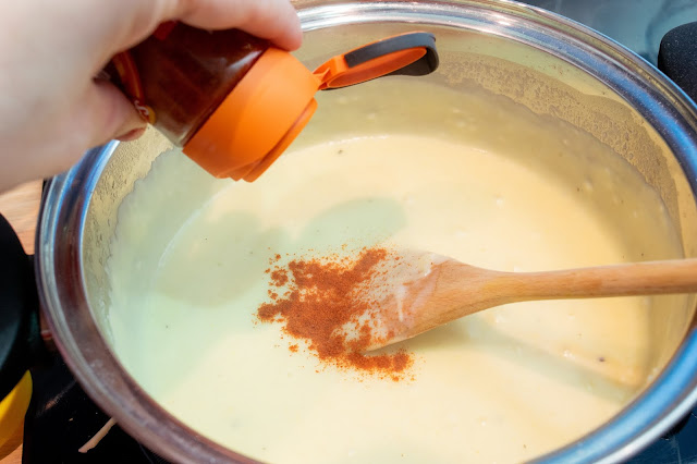 A bottle of smoked paprika being sprinkled into a creamy sauce in a metallic silver pan.