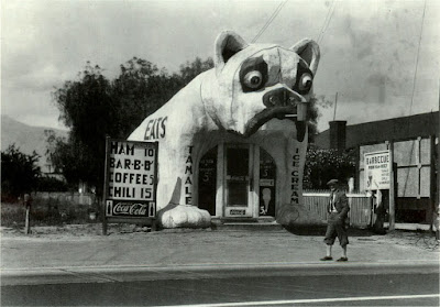 A black-and-white photograph of a restaurant shaped like a bulldog. "TAMALE" is written on the bulldog's left leg. "ICE CREAM" is written on the bulldog's right leg. A man walks down the street in the foreground.