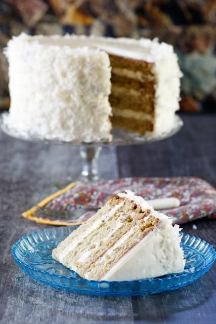 Four layers of moist banana cake wrapped in fluffy marshmallow like meringue frosting is bound to make a statement. Add a little coconut to mix to make it extra special and this star-spangled cake is perfect for a party! layered banana cake on stand with slice of cake served in front of it