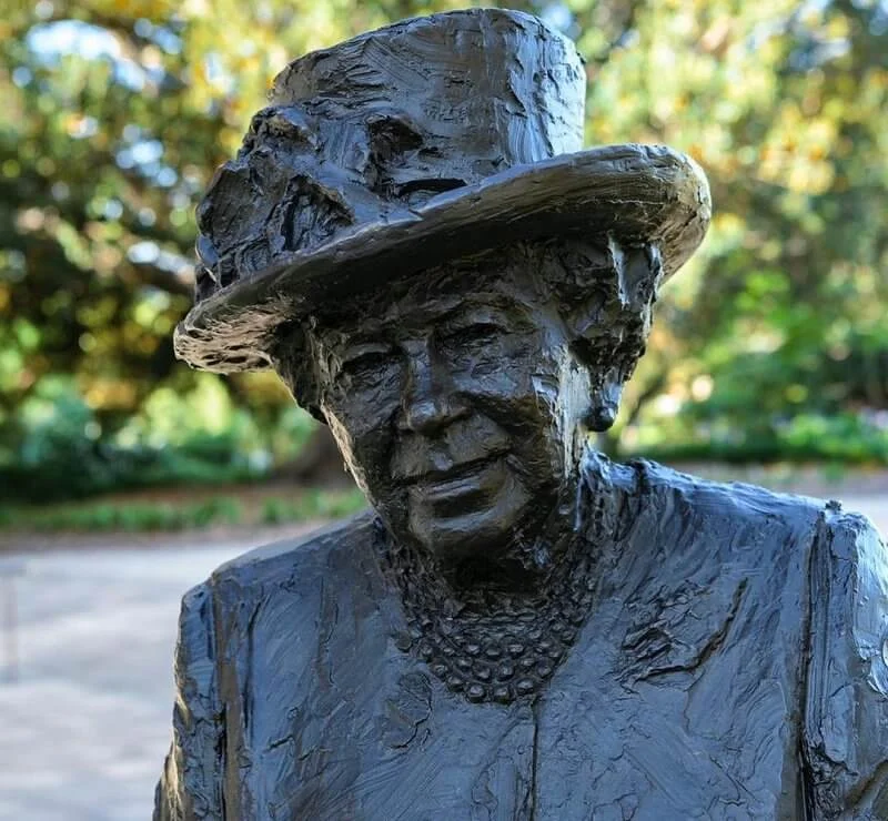 The sculpture depicts Queen Elizabeth in a coat and hat carrying her trademark Launer handbag. diamond silver brooch and pink dress