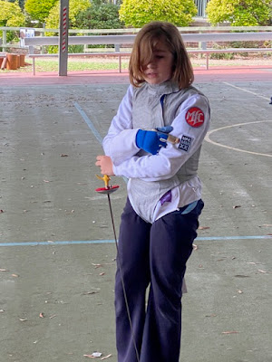 Jemima stands in the playground, wearing long black pants, cyan sneakers, white fencing jacket and foil lamé. She holds a fencing foil, pointing down, in her left hand. She wears a fencing glove on her right hand and points to the patches sewn to her left sleeve. The top one is a round orange one with purple border and MFC in white, below it is a rectangular black one with a pewter border and MFC, MM, XX stacked.