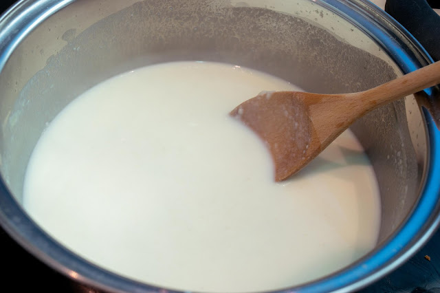 A creamy mixture of milk, butter and flour in a metallic silver pan with a wooden spoon sitting on the side.