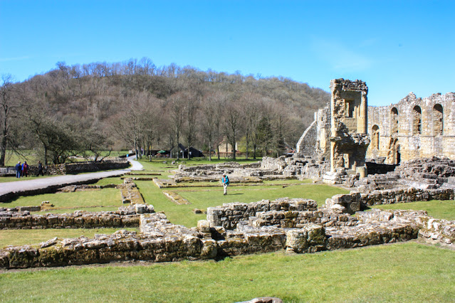 A fairly flat grass area with large ruins on the right but lots of small walls on the ground that indicate where rooms once were.
