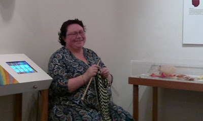 Between the display cabinets sits a woman (Cheryl) knitting with large wooden needles and a stripy fabric. 