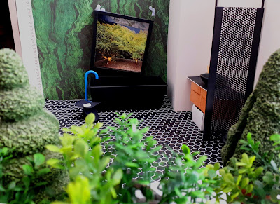 One-twelfth scale modern miniature half-built scene of a modern bathroom with green marble walls, black tiled floor and black fittings. On the wall above the bath is a window, falling down showing the Blutak that held it, and in the front if the picture is a selection of miniature plants.