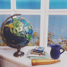1/12 miniature scene of a windowsill overlooking the sea. On the windowsill is a metal globe, a stack of old books, a blue jug and an old telescope.
