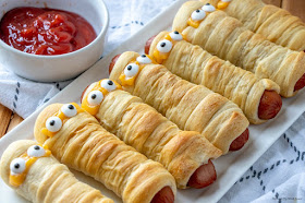 Hot dogs wrapped in crescent rolls with eyes on white platter with a ketchup on the side