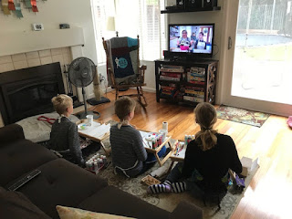 Three children sitting on the floor in a living room, watching a person on a television.