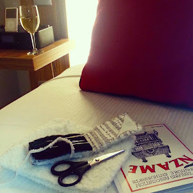 Bed in a hotel room with an NZAME magazine, a finished piece of knitting, scissors and a needle case on it. On the bedside table is a glass of wine and an iPod.