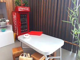 1/2 scale modern miniature asian cafe with a counter, drinks fridge, rustic fence with a bamboo plant in front of it and a metal table with a bowl of soup on it and wooden stool and a back pack in front of it.