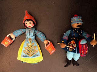 two felt dolls- one wearing a yellow & blue dress, holding two buckets, and the other wearing a blue uniform and a red drum