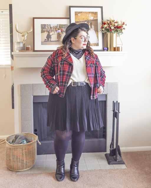 An outfit consisting of a black wide brim hat, black and white striped turtleneck under a white sweater half tucked unto a black pleated mini skirt under a plaid cropped moto jacket and black chelsea boots.