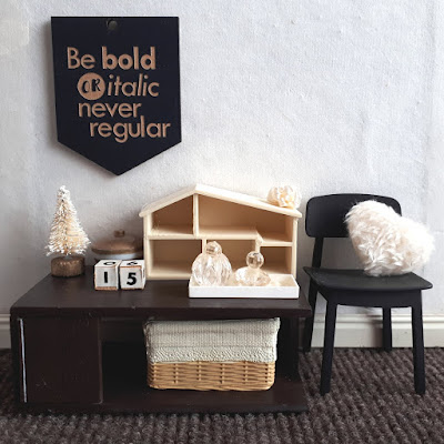 One twelfth scale modern miniature cupboard-top scene in cream and brown containing a tiny bottlebrush Christmas tree, a lidded bowl with gold and white stripes, wooden blocks with the numbers '1' and '5' on them, and an empty dolls' house. Above it on the wall is a wallhanging that says 'Be bold or italic, never regular' and to the right, a black wooden chair with a cream fluffy heart-shaped cushion on it.