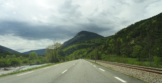 Highway mit rechts Bahngleisen, links einem Fluß, im Hintergrund Berge und grüne Wälder, leere Fahrbahn