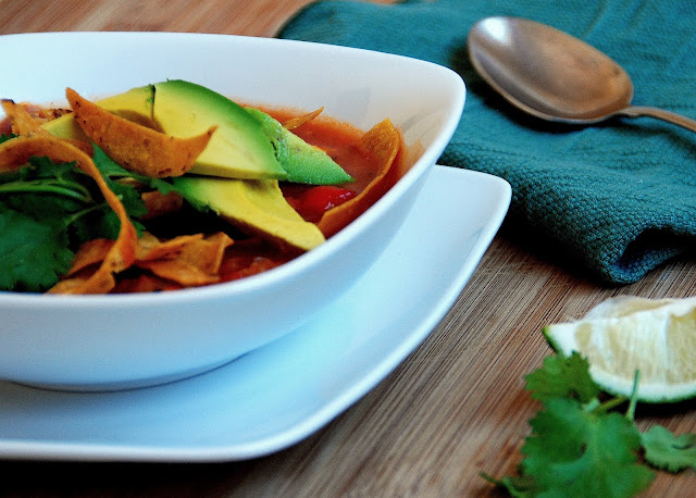 tortilla soup topped with sliced avocado, tortilla strips, and cilantro in white bowl on white plate in front of a spoon on a blue napkin