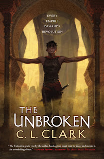 A Black woman with short hair, wearing a leather sleeveless tunic and leather pants with a red sash and a long dagger at her belt stands with her arms braced against the pillars of a large keyhole-shaped doorway, the light is golden behind her and dust rises at her feet.
