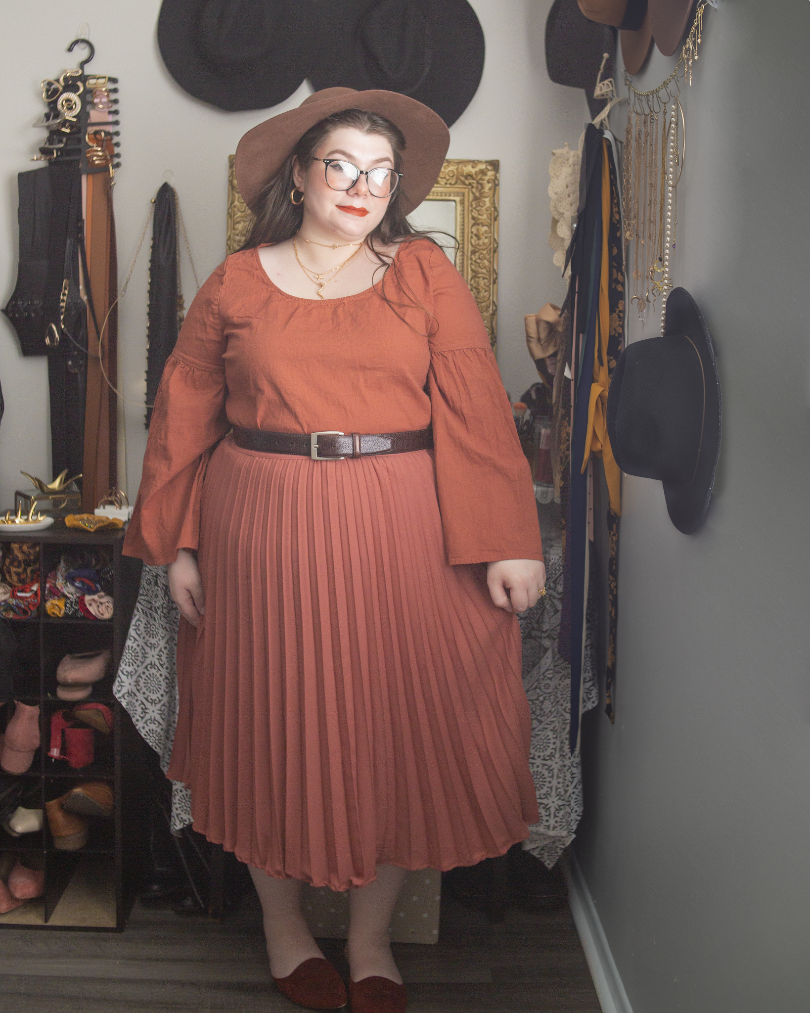 An outfit consisting of a reddish brown wide brim floppy hat, a burnt orange bell sleeve linen dress, worn as a top, tucked under a rosey brown pleated midi skirt and burnt orange pointed toe mules.