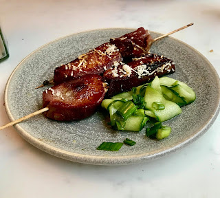 Two thin light brown cylindrical skewers filled with dark brown ox tongue with green ribbons of cucumber at the side of it on a circular blue plate on a light background 