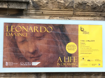 Photo of poster outside museum, showing a da vinci sketch of a a woman's face and yellow  text reading 'Leonardo da Vinci, a life in drawing'
