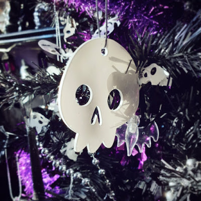 White skull decoration A cartoonish white acrylic skull, very glossy, hanging in the black Christmas tree, with a clear bat-shaped LED string light, silver bead garland, purple tinsel, and black tinsel with white skulls. The shelve and lantern-styled string lights are visible in the background, out of focus, in the upper left