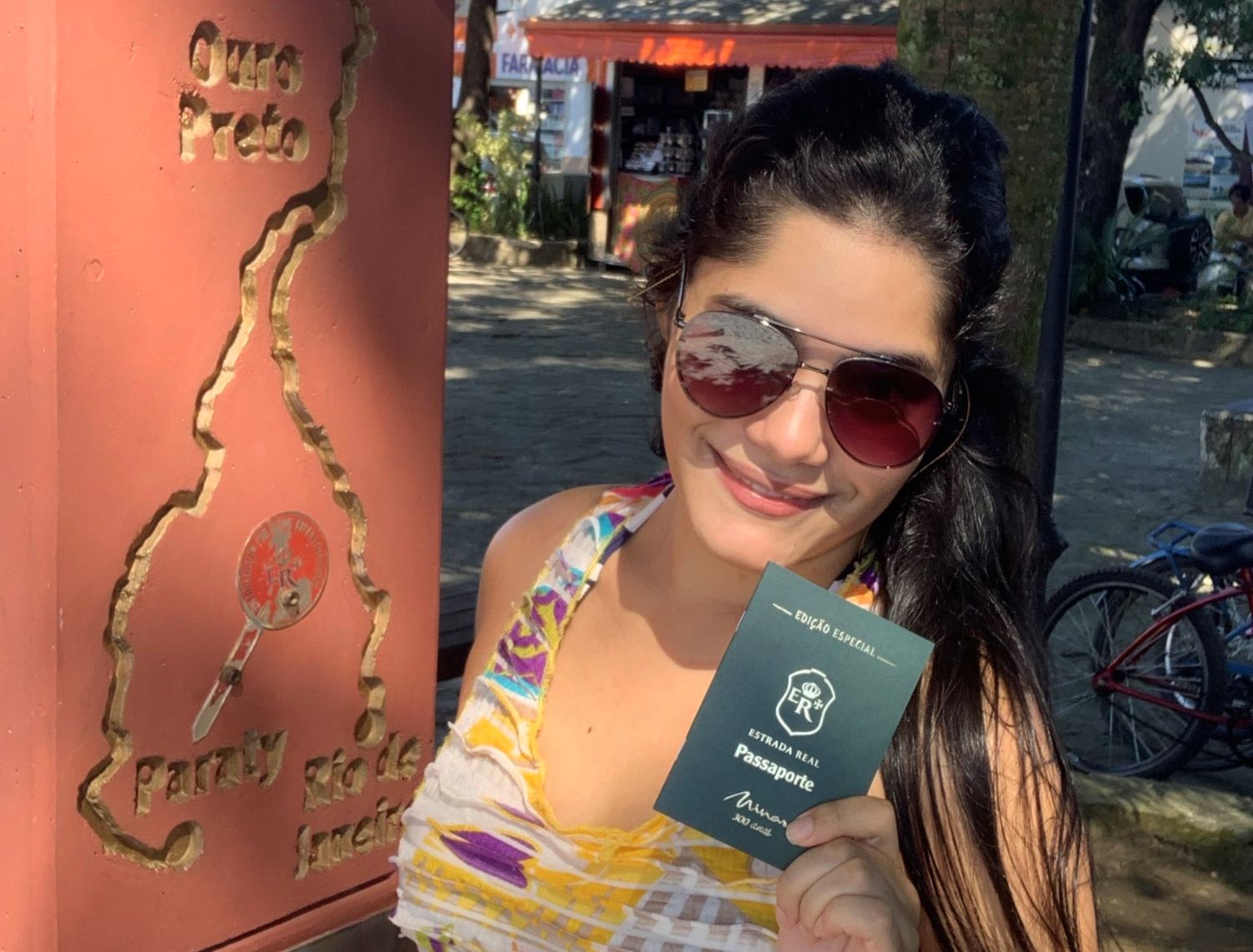 passaporte estrada real em paraty mulher jovem, branca, de cabelos pretos e oculos escuro, segurando um passaporte verde com estrada real escrito na capa