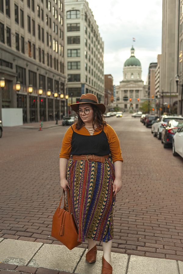 An outfit consisting of a brown panama hat, a burnt yellowish orange lace up 3/4 sleeve shirt layerd under a black camisole dress tucked under a warm patterned maxi skirt and brown d'orsay flats.