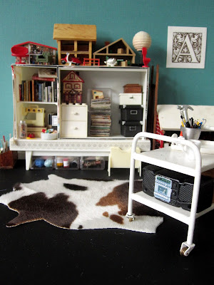 1/12 scale miniature studio with a trolley in the foreground holding a boom box and various tools, and a shelving unit in the background with storage boxes, books, magazines, a sewing machine and miniatures.