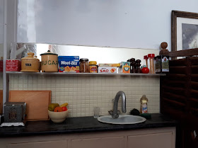 1/12 scale modern miniature scene of a kitchen bench with a fold-down toaster, chopping board and bowl of fruit on it. Next to the sink is a glass, a scourer, a sponge and a bottle of washing-up liquid. On the shelf above are two containers of tea, a cannister of sugar, a packet of Weet-Bix, a half-used jar of jam, a jar of vegenite, a packet of Sao crackers and a packet of Lemon crisp biscuits, a box of barley sugars, a salt and pepper grinder, a tomato-shaped tomato sauce bottle, a jar of spaghetti and a cask of red wine.