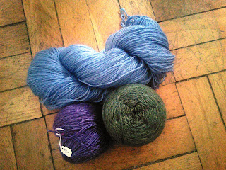 a twisted skein of sky blue yarn, a yarn cake of purple yarn, and a yarn cake of dark green yarn