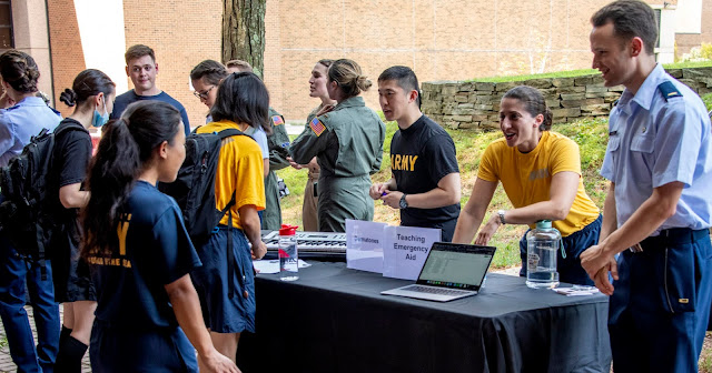 A student standing in front of a table at the Connections Fair, with other students around.