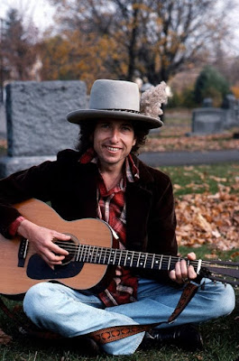Bob Dylan ca 1975 color photo sitting on the ground, wearing a hat with plumage and playing a guitar