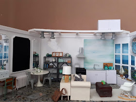 1/12 scale modern miniature scene of a kitchen, dining room and lounge in shades of white, teal, grey and light wood in a flat overlooking the sea.