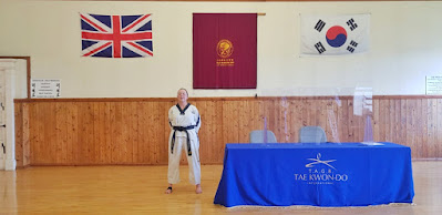 Very sweaty 51 year old lady in a Tae Kwon-Do suit stands in a hall next to a table draped with TKD logos, behind on the wall are TKD, GB and South Korean flags.