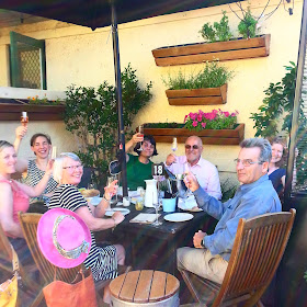 Group of people around an outdoor table, toasting the photographer with glasses of sparking wine.