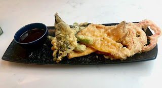 A selection of light brown shaped tempura pieces next to a black pot filled with dark brown liquid on a rectangular black plate on a bright background