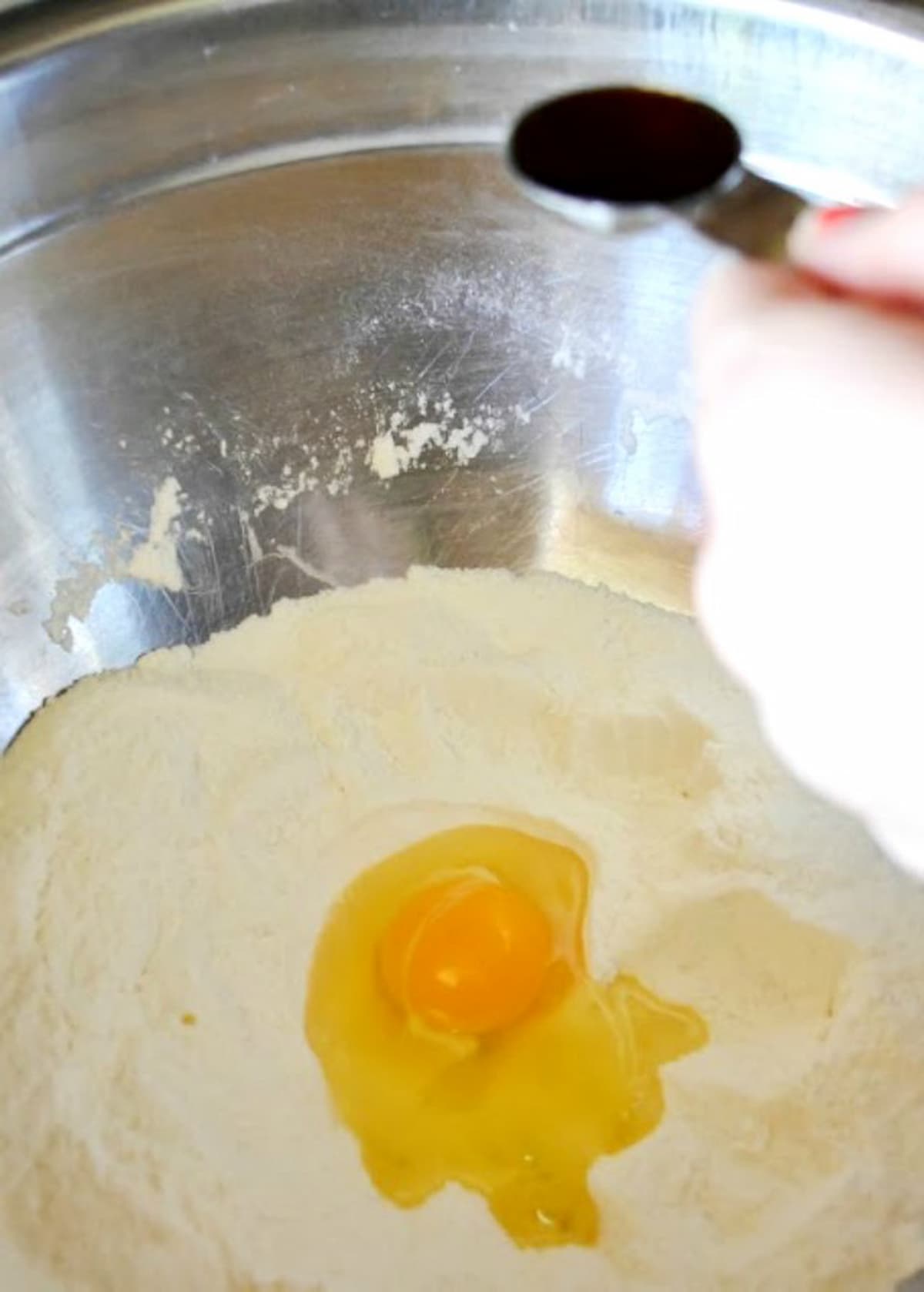 Flour, Sugar and Baking Powder mixed together in a mixing bowl with an egg in the center and a teaspoon of Vanilla being held over the top of bowl.