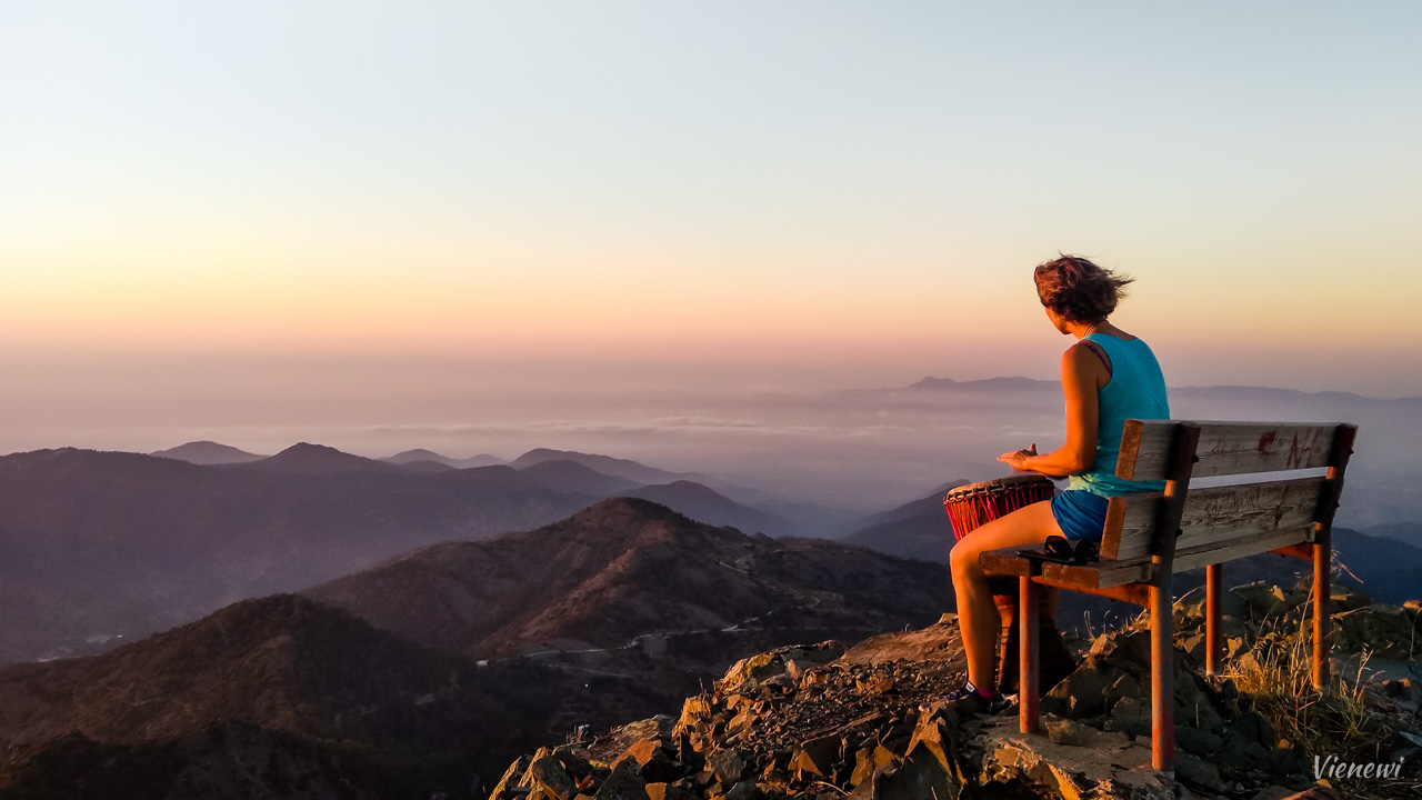Pejzaż widziany z punktu widokowego w kierunku północno zachodnim. Z lewej strony kadru kobieta siedząca na ławeczce i grająca djumbe i w tle szczyty gór spowite chmurami i oświetlone zachodzącym słońcem.