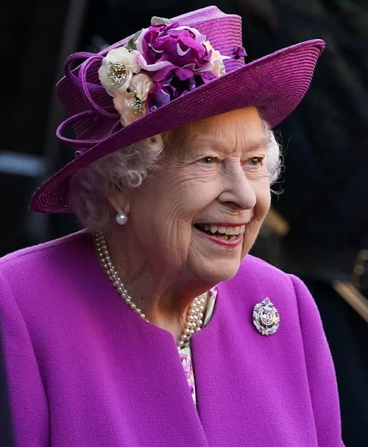 The Queen wore a floral print dress and purple coat and purple hat. diamond brooch