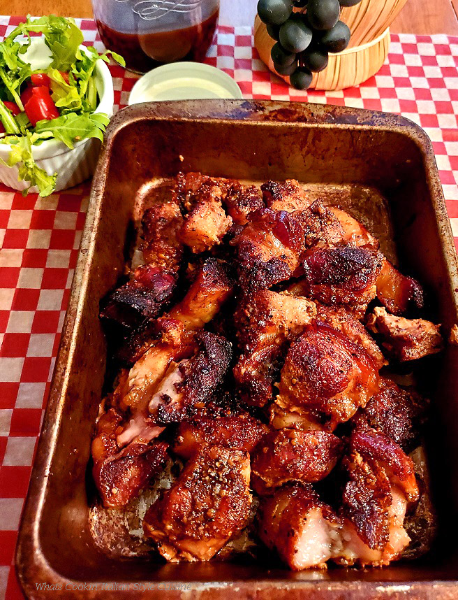 This is an old roasting pan that has pork cubed made into burnt ends. It is glazed with a sweet shiny apricot, barbecue hot sauce and has a arugula salad with tomato on the side. This pan sits on a checkerboard napkin style paper